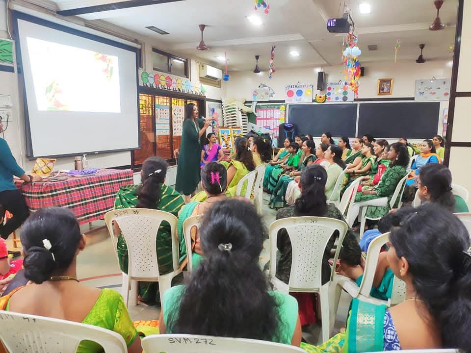 A facilitator speaking with a microphone to a hall full of women, a slide projected beside her